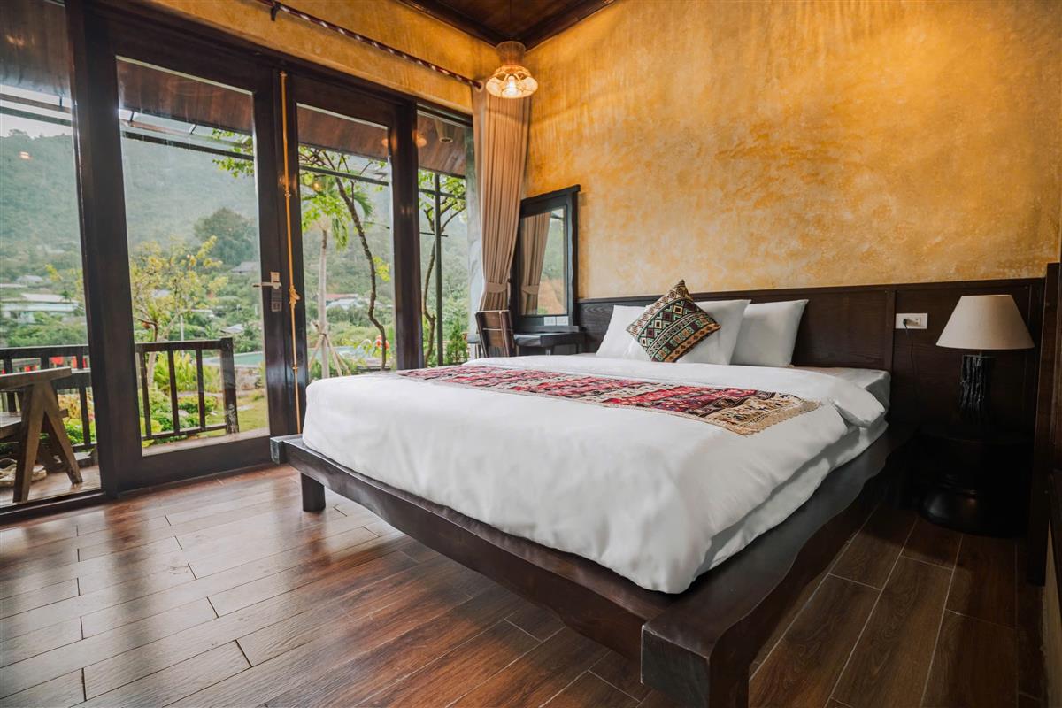 Phòng ngủ tại Mít Retreat với nội thất gỗ giản dị, cửa kính lớn và ban công nhìn ra thiên nhiên xanh mát Phòng ngủ tại Mít Retreat với nội thất gỗ giản dị, cửa kính lớn và ban công nhìn ra thiên nhiên xanh mát